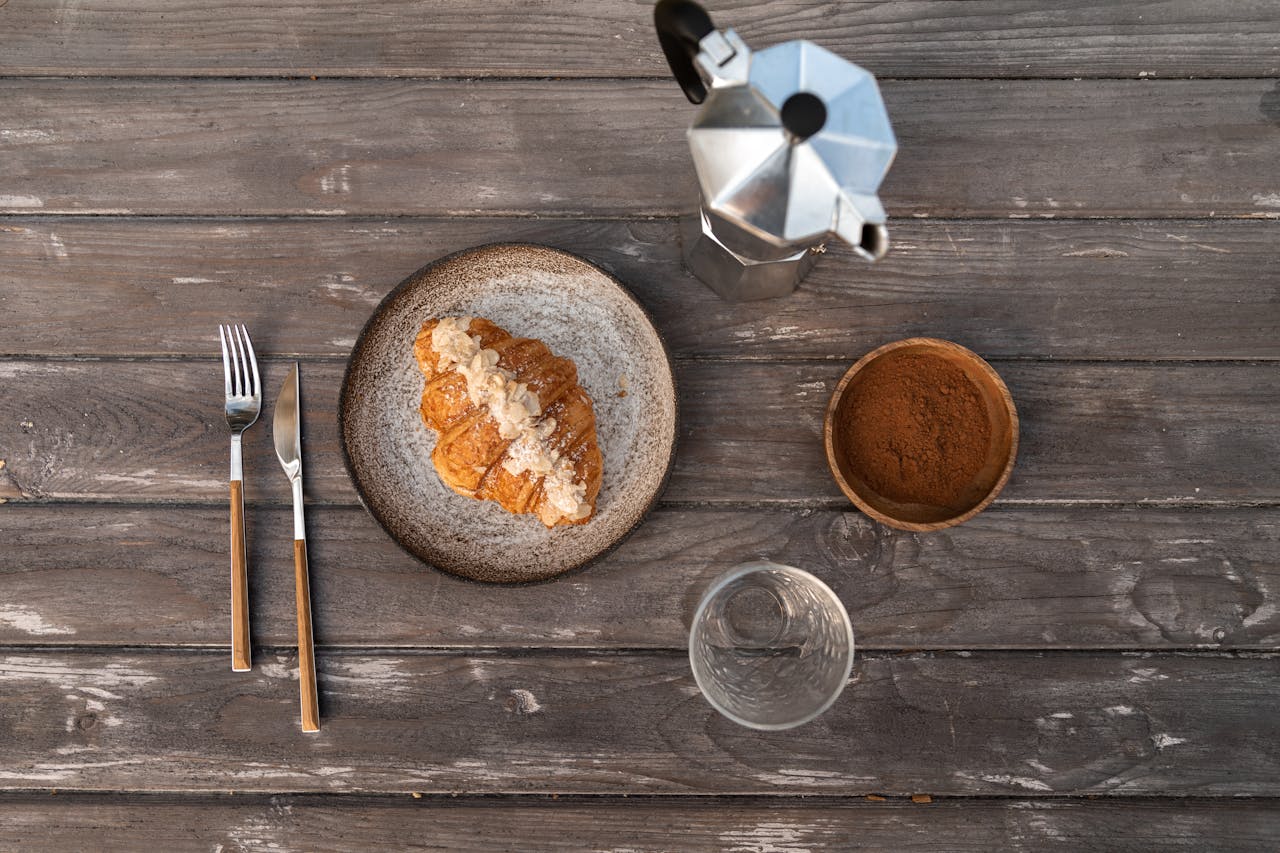 about-img Flat lay of a croissant on a plate with a moka pot and coffee grounds on wooden table.