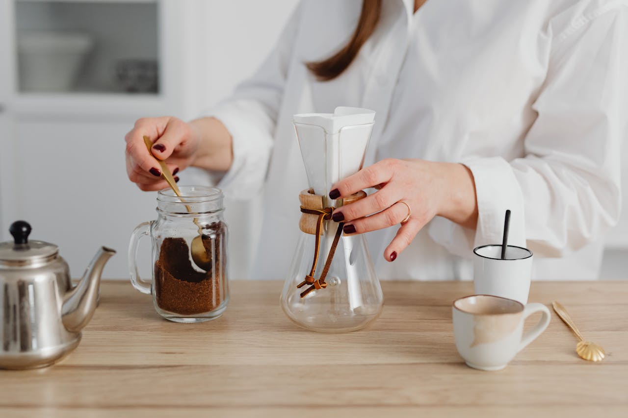 contact-img Hands preparing coffee using a Chemex and jar on a wooden surface, highlighting the brewing process.