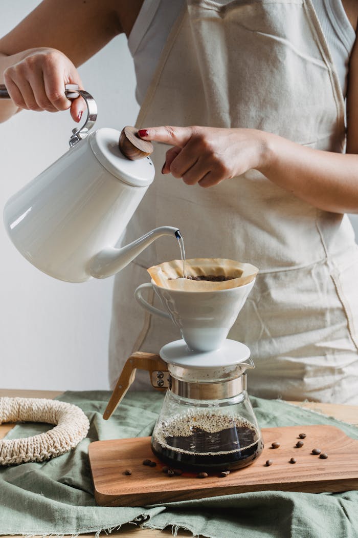 services-02 Woman preparing pour over coffee with a white kettle and dripper in a cozy kitchen setting.